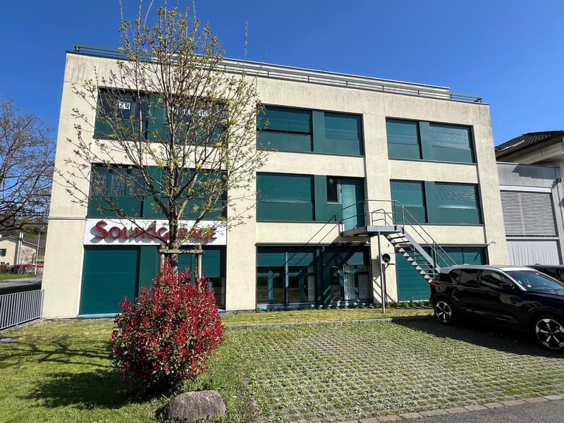 A multi-story commercial building with a green and white exterior. The building has large windows and a prominent sign that says 'Soundcraft'. There is a paved parking area in front of the building, and some landscaping with a blooming tree and bushes.