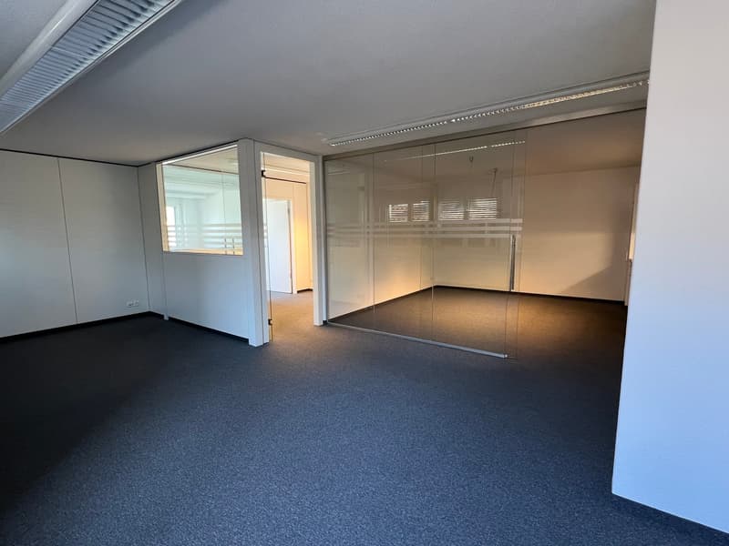 The image shows a large, open office space with gray walls, a gray carpet floor, and glass partitions. The space appears to be a commercial or professional office environment, with a clean and modern design.
