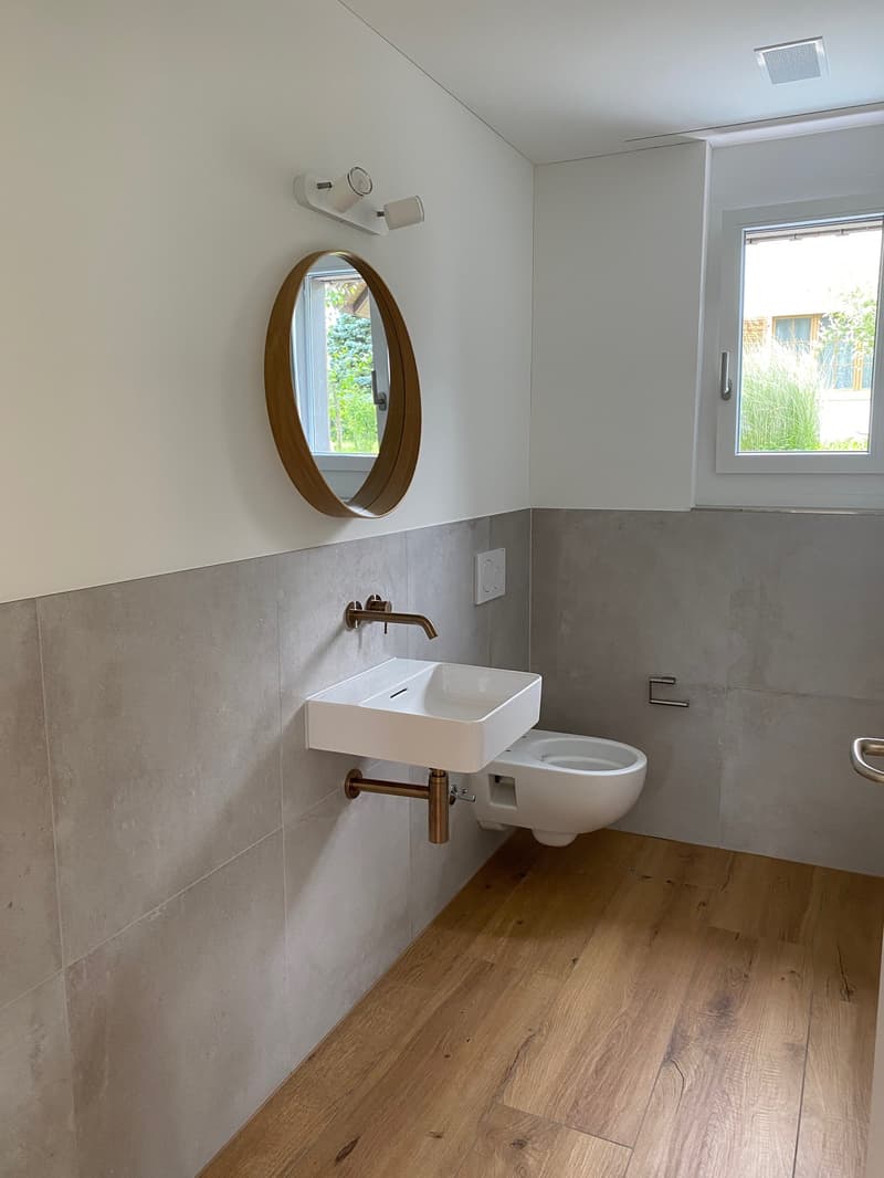 The image shows a modern bathroom with a white sink, a round wooden mirror, and a toilet. The floor is made of wood, and the walls are tiled in a light gray color. The bathroom has a minimalist and clean design.