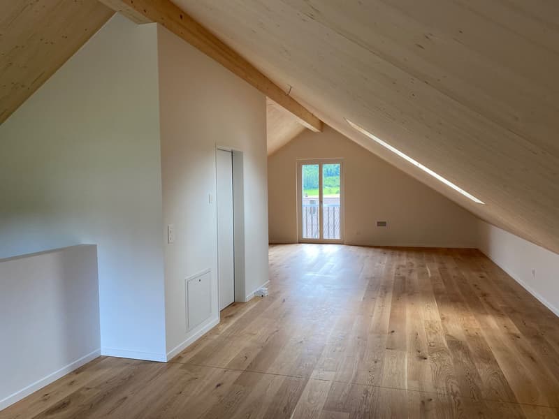 Spacious attic room with slanted wooden ceiling, hardwood flooring, and a large window providing natural light. The room appears to be empty and ready for furnishing.