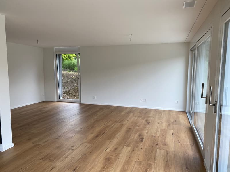 Spacious living room with large windows, hardwood flooring, and access to a balcony or terrace overlooking a green outdoor area.