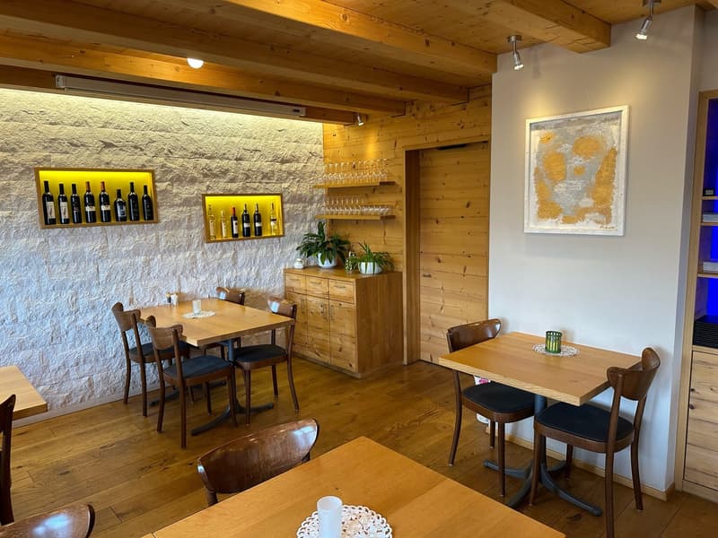 The image shows a cozy and rustic interior of a restaurant or cafe. The walls are made of exposed stone and the ceiling has wooden beams, creating a warm and inviting atmosphere. There are several wooden tables and chairs, as well as a wooden cabinet disp