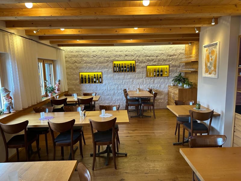 The image shows a cozy and rustic-style restaurant or cafe interior. The space features wooden beams on the ceiling, a stone accent wall with shelves displaying wine bottles, wooden tables and chairs, and a warm, inviting atmosphere.