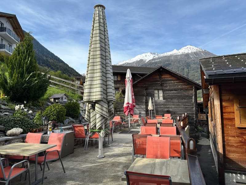 Outdoor seating area with wooden tables and chairs, large striped umbrella, surrounded by wooden buildings and mountains in the background