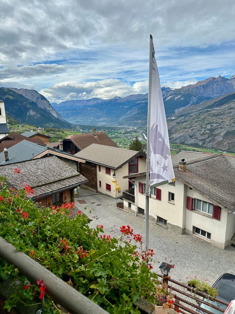 The image shows a picturesque mountain village with traditional Swiss-style buildings, surrounded by lush greenery and set against a backdrop of majestic snow-capped peaks. A large white flag with stars is prominently displayed, adding to the scenic charm