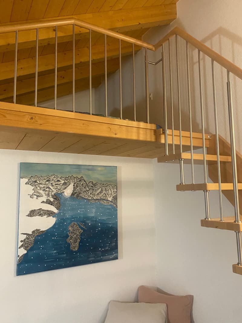 The image shows a cozy interior with a wooden ceiling and staircase. The staircase has a metal railing, and there is a painting of a coastal landscape on the wall. The room appears to have a balcony or terrace, as indicated by the visible feature.