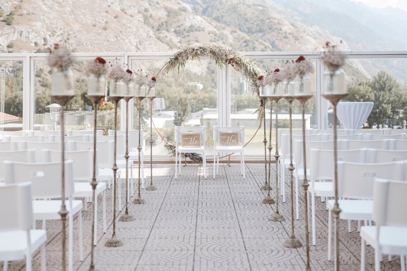 Outdoor wedding ceremony setup with a floral arch, white chairs, and a scenic mountain view in the background. The aisle is decorated with a patterned floor and candle holders lining the sides.