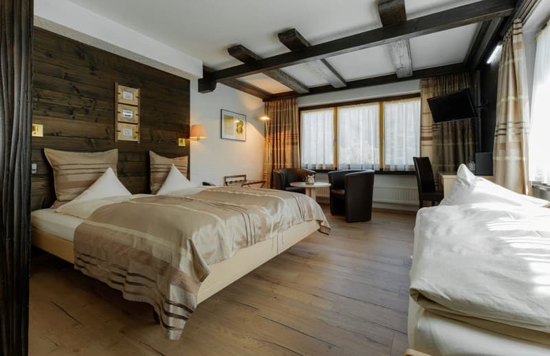 Spacious bedroom with wooden walls and ceiling beams, large windows, hardwood flooring, king-size bed with beige bedding, armchair, and a fireplace