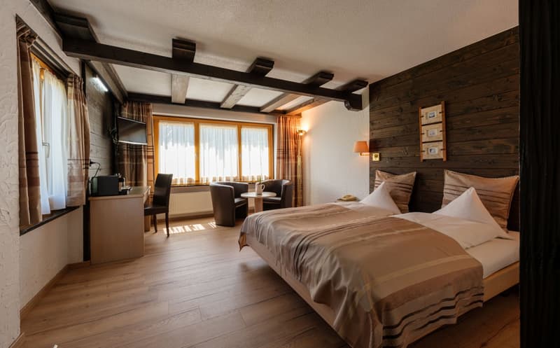 Spacious bedroom with wooden beams on the ceiling, wooden wall paneling, large windows, a seating area with a table and chairs, and a king-size bed with neutral-colored bedding.