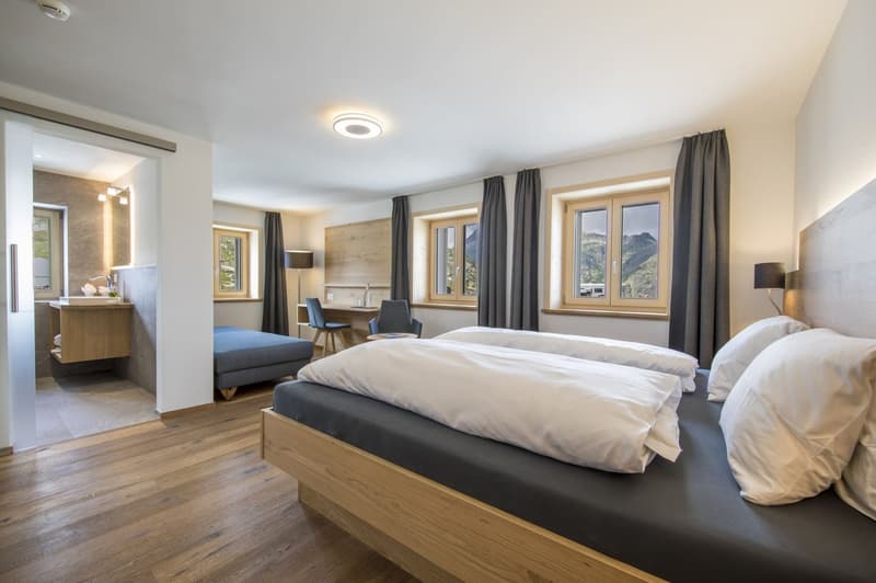 Spacious bedroom with large windows offering a scenic mountain view, wooden flooring, a king-size bed, a seating area, and an en-suite bathroom