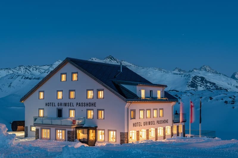 A large, multi-story hotel building with a balcony and terrace, located in a snowy, mountainous landscape with a clear blue sky. The hotel is illuminated at night, with several lit windows visible.