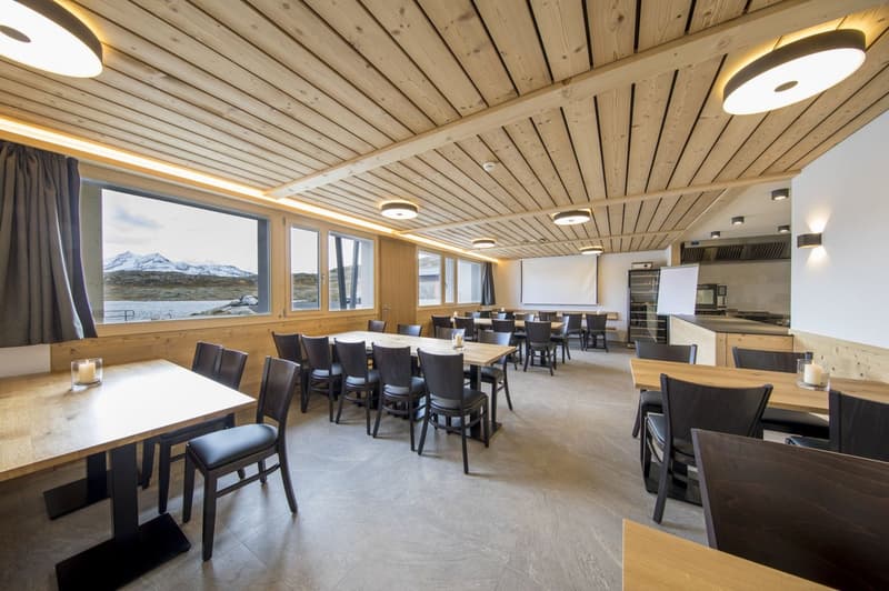 Large dining room with wooden ceiling and walls, large windows overlooking a scenic mountain landscape, multiple dining tables and chairs, kitchen area visible in the background
