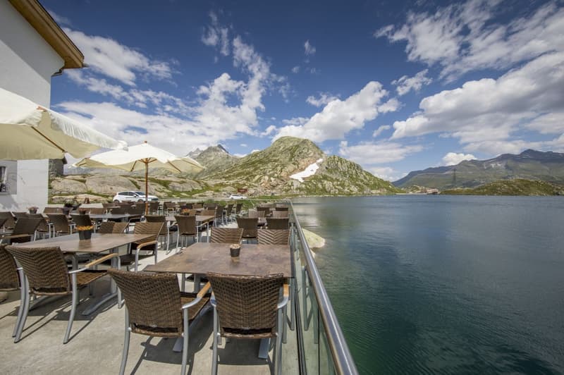 Outdoor restaurant terrace overlooking a scenic lake surrounded by mountains, with tables and chairs set up for dining with a beautiful view of the landscape.