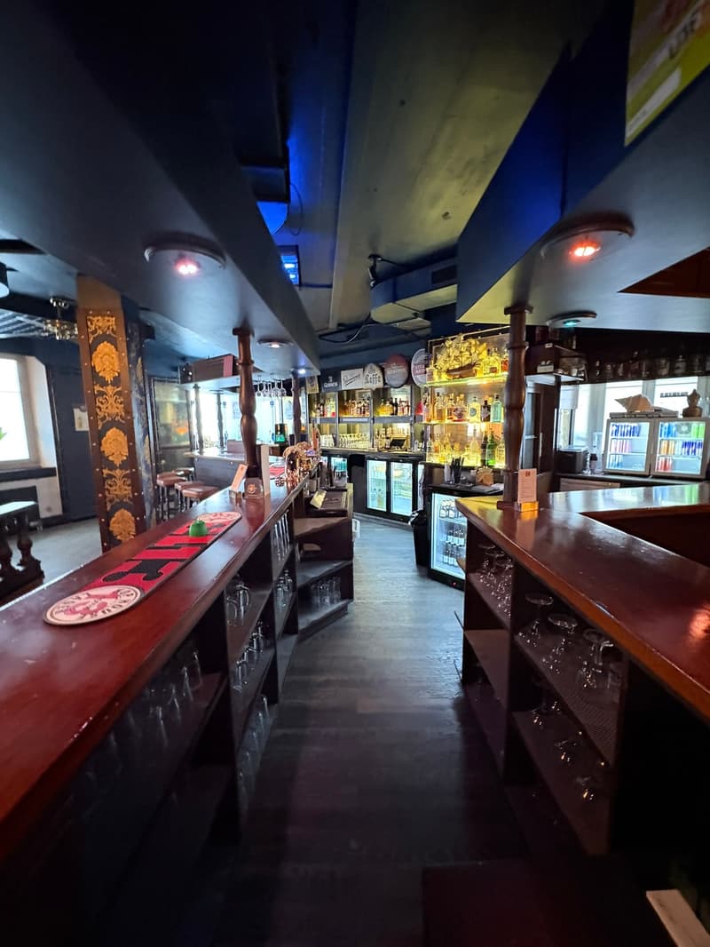 A bar interior, with red countertops, shelves with glasses and bottles, wooden furniture, illuminated ceiling lights, and a blue-colored ceiling.