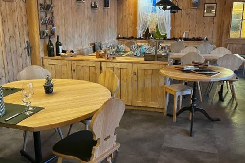 Round tables with chairs, wooden cupboards, a counter, wine bottles, and a picture frame are arranged in a restaurant.