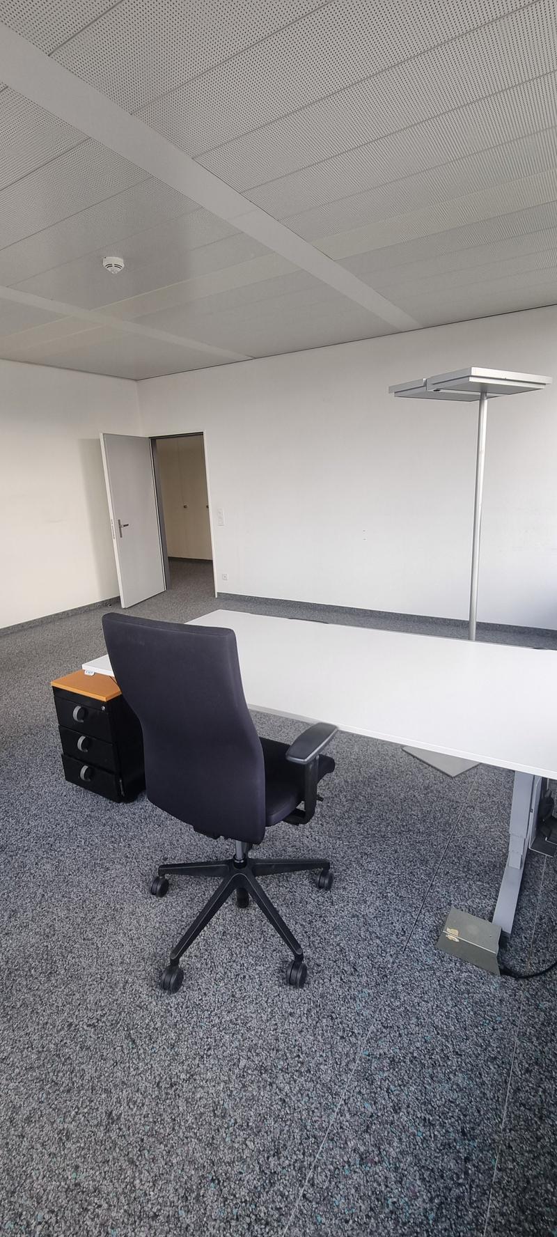 The image shows an empty office space with a desk, an office chair, and a floor lamp. The room has a gray carpet floor and a suspended ceiling with a grid pattern. The walls are white, and there is a door visible in the background.