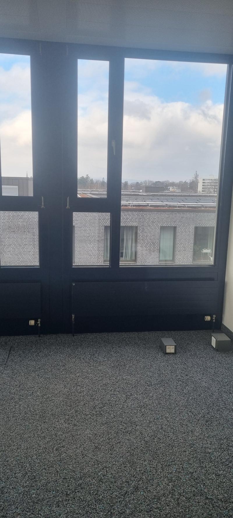 Large windows with a view of the cloudy sky and buildings outside, dark-colored walls, gray carpeted floor, and a small electrical device on the floor.