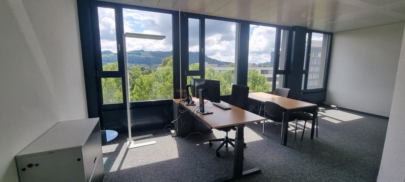 Large office space with floor-to-ceiling windows offering a scenic view of the surrounding landscape. The room features a long wooden desk with multiple computer monitors, chairs, and other office equipment. The space appears to be well-lit and modern in