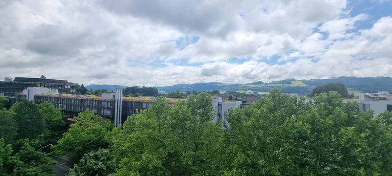 The image shows a panoramic view of a city surrounded by lush green trees and hills. The cityscape includes a mix of modern and older buildings, with a cloudy sky overhead. The landscape appears to be in a mountainous or hilly region, providing a scenic b