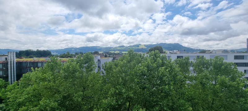 The image shows a panoramic view of a city surrounded by lush greenery and rolling hills. The cityscape is dominated by modern multi-story buildings and apartments, with a mix of residential and commercial structures. The sky is partly cloudy, creating a