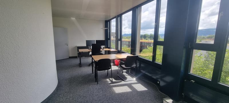 Large open office space with floor-to-ceiling windows providing a scenic view of the surrounding landscape. The room is furnished with multiple desks, chairs, and computer equipment, suggesting it is used as a workspace or office. The overall design appea