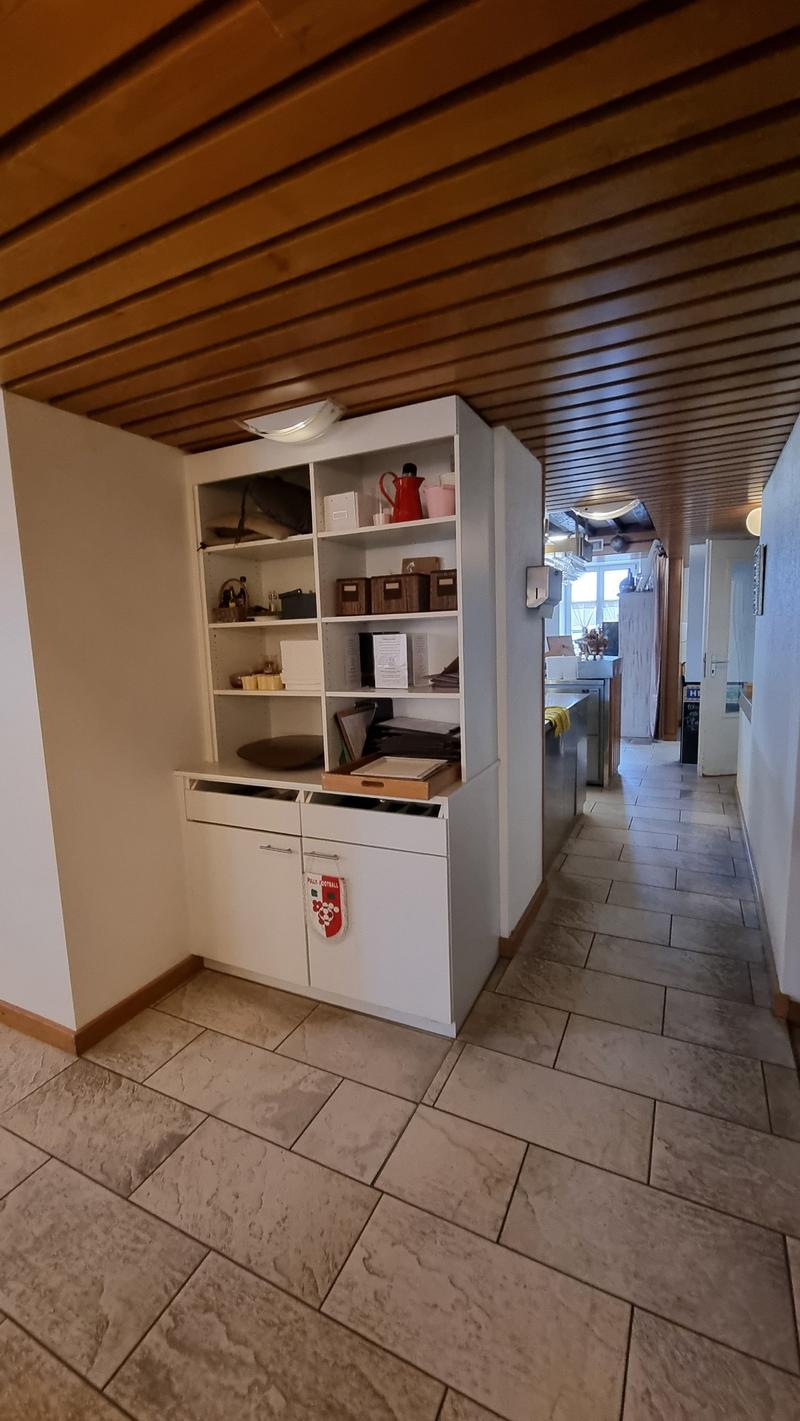 A room with a white cabinet, tiled floor, and wooden ceiling. The cabinet has drawers and shelves with various items on them. The room has a door on the right side, and a window on the left side. The room is empty, and there is a light on the ceiling.