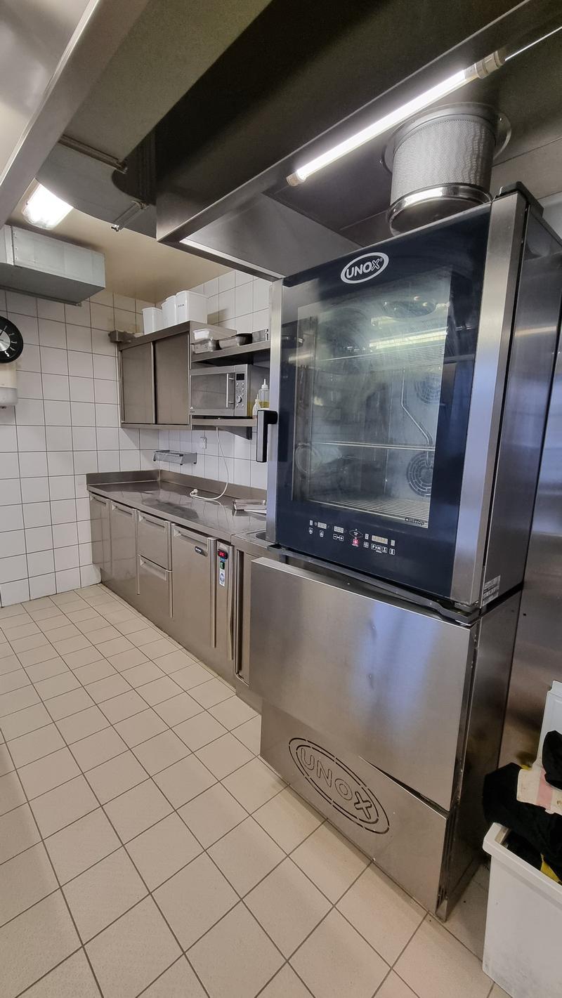 Commercial kitchen with stainless steel appliances, tiled floor, microwave, oven, sink, cabinets, and a large UNOX oven.