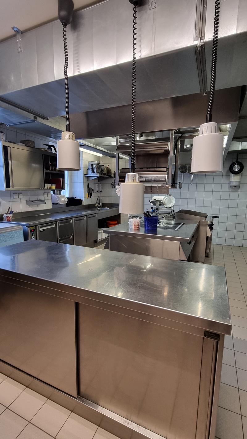 Commercial kitchen with stainless steel countertops, hanging lamps, tiled walls, and various kitchen appliances and utensils.