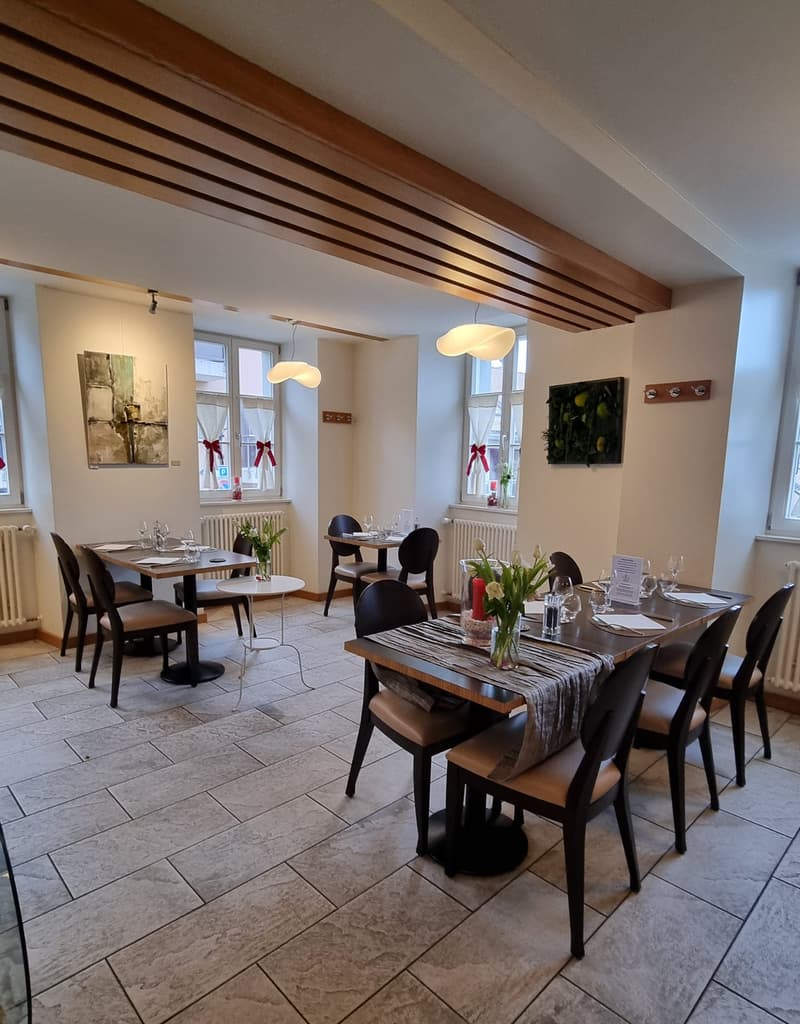 A restaurant with wooden ceiling, white walls, tiled floor, tables and chairs, radiators, and paintings on the wall.