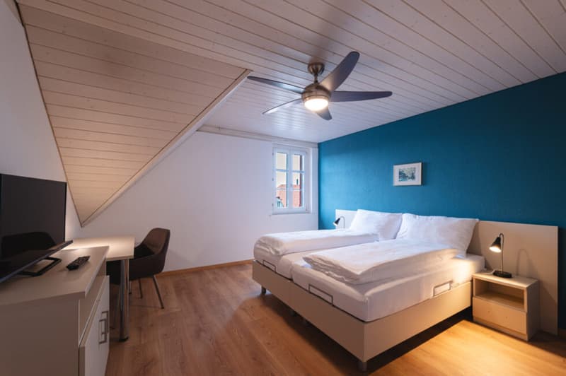 Bedroom with two beds, desk, chair, TV, lamps, wooden floor, blue wall, ceiling fan
