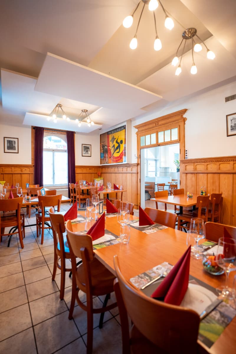 Wooden walls, wooden tables and chairs, multiple chandeliers, picture frames on the wall, red napkins on the table