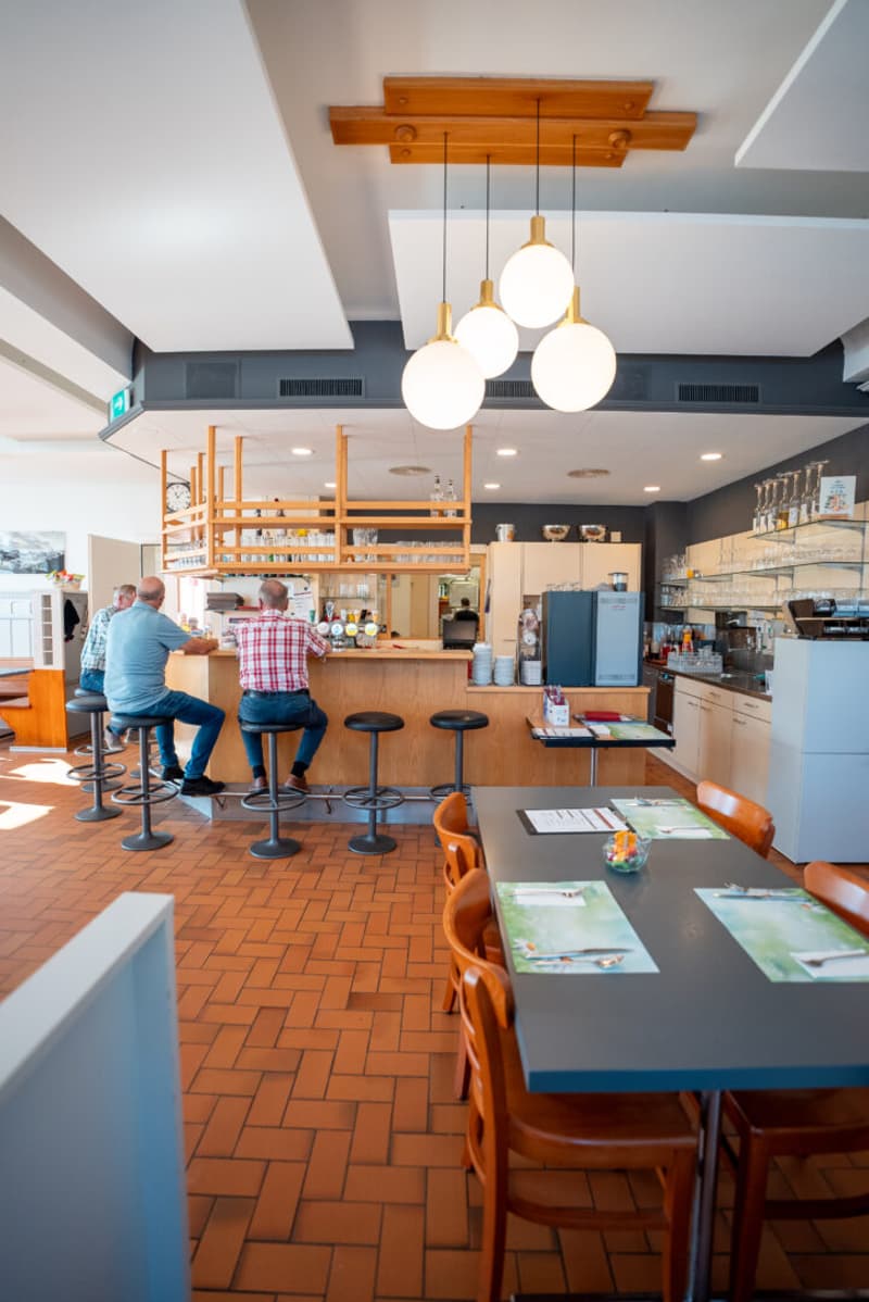 A restaurant with a bar counter, stools, tables, chairs, and a wooden shelf with glasses.