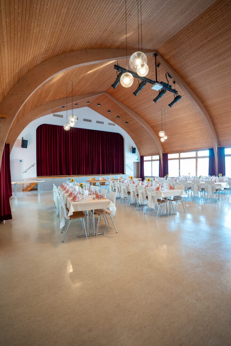 large room, wooden ceiling, tables and chairs, stage with red curtain, windows