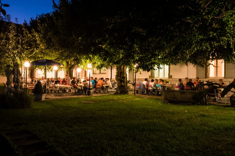Outdoor seating area with tables and chairs, umbrellas, trees, and a building in the background.