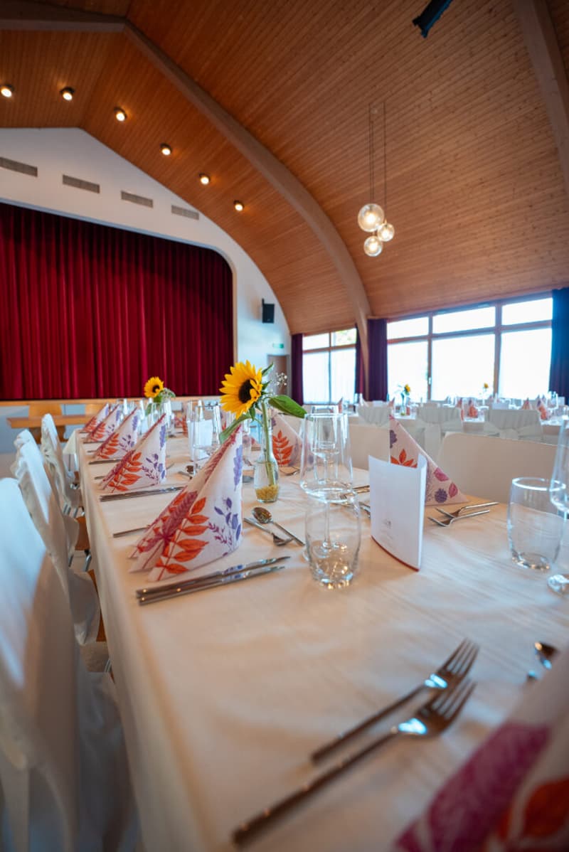 Dining room with long table, white chairs, white tablecloth, sunflowers, glasses, cutlery, napkins, and a red curtain