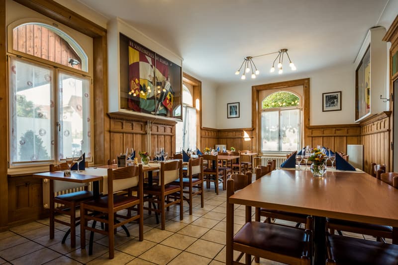 Dining room with wooden tables and chairs, white tiled floor, wooden walls, large windows, chandelier, paintings, radiators, flowers in vases
