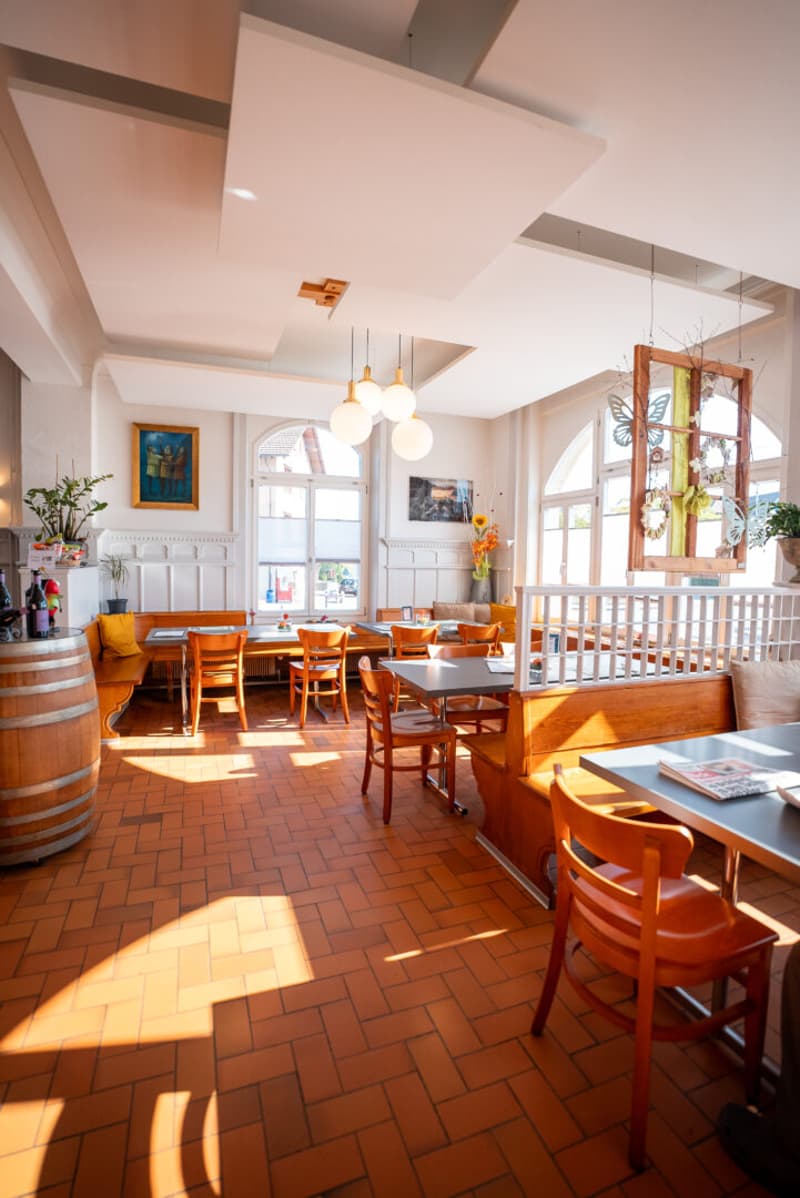 A restaurant with wooden tables and chairs, a wooden barrel, a white ceiling, and a white wall with a picture frame. There are also lamps hanging from the ceiling, a potted plant, and a vase with flowers on the table. The restaurant has a view of the outs