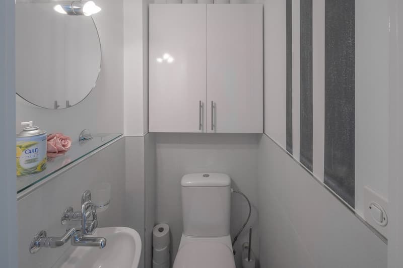 modern bathroom with white tiles, white sink, white toilet, white cabinets, mirror, and a glass shelf