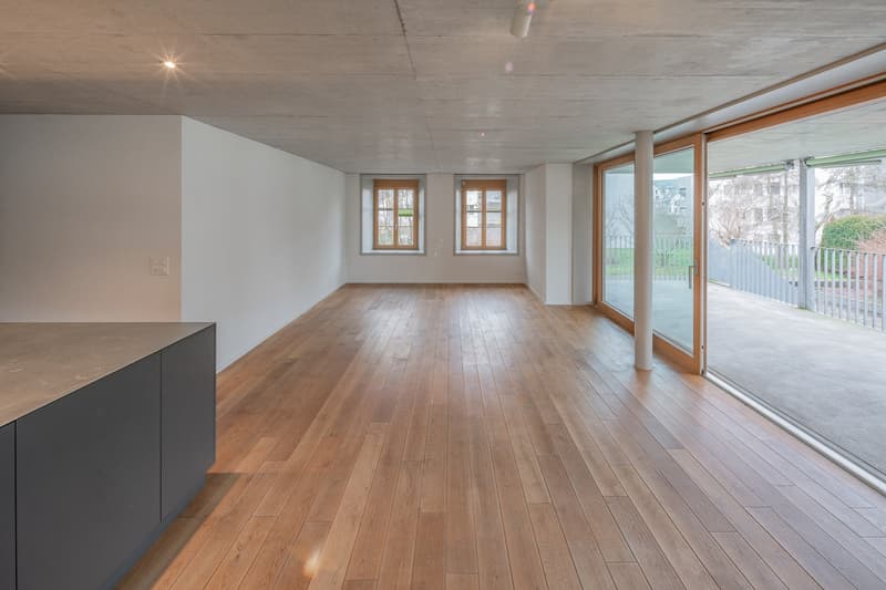 An empty room with hardwood floors, white walls, glass windows, glass doors leading to a balcony, and recessed lighting.