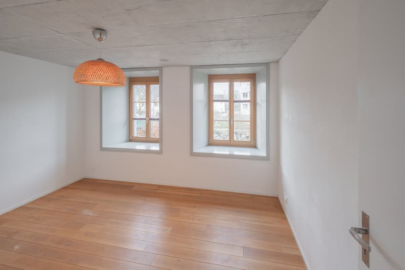 Empty room with wooden floors, white walls, two windows, and a pendant lamp.