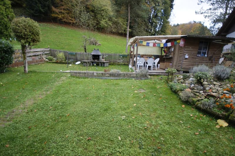 Open grassy space with wooden fence, small porch with chairs, outdoor fireplace, colorful flags
