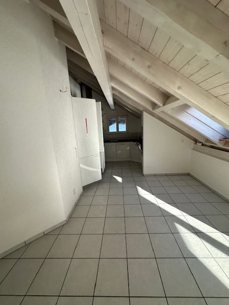 an attic room with white tiled floor, wooden beams, an open door, and a kitchen area