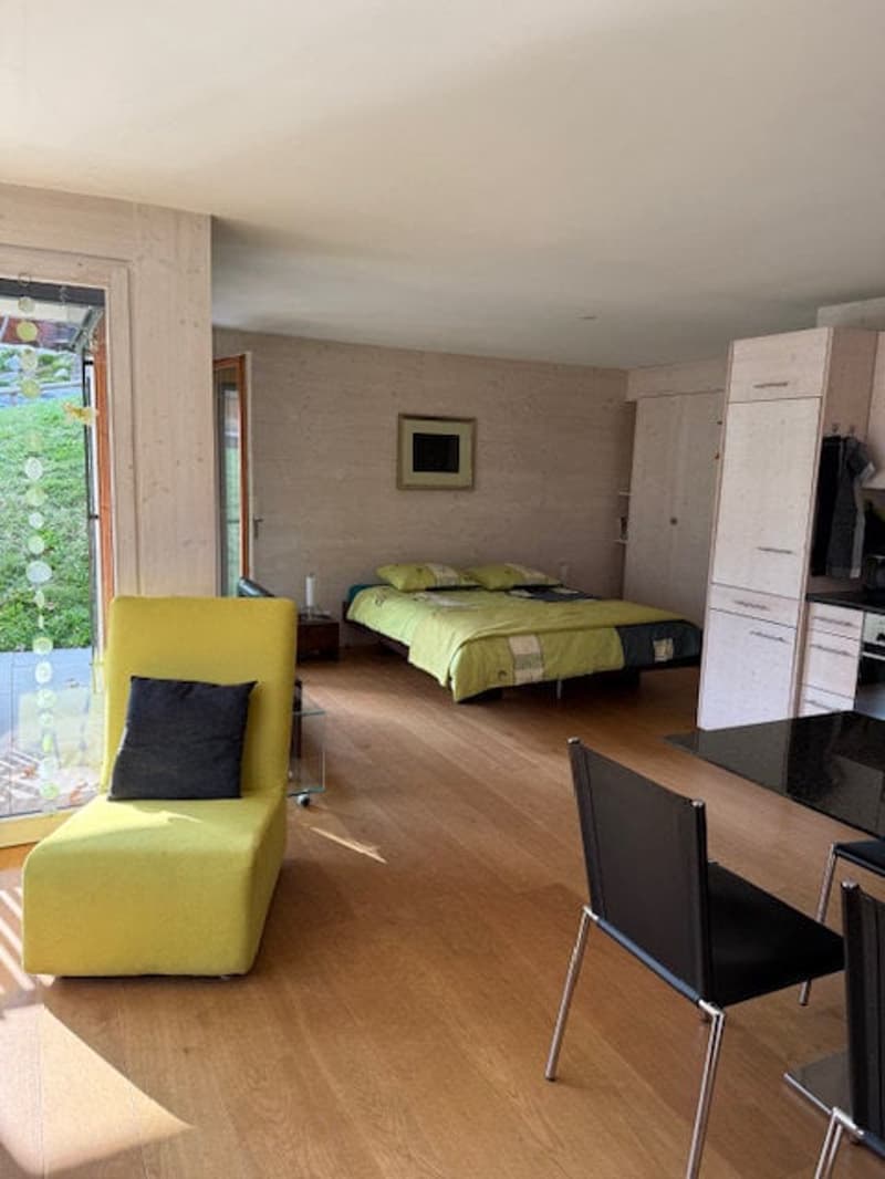 A bedroom with a bed, a yellow chair, a table, and a kitchen area with cabinets and a countertop