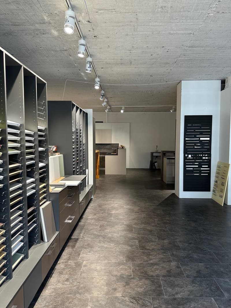 modern showroom with open floor plan, minimalist design, ceiling track lights, tiled floor, high shelves with samples, contemporary cabinets, open kitchen area with countertops and island, light and spacious layout