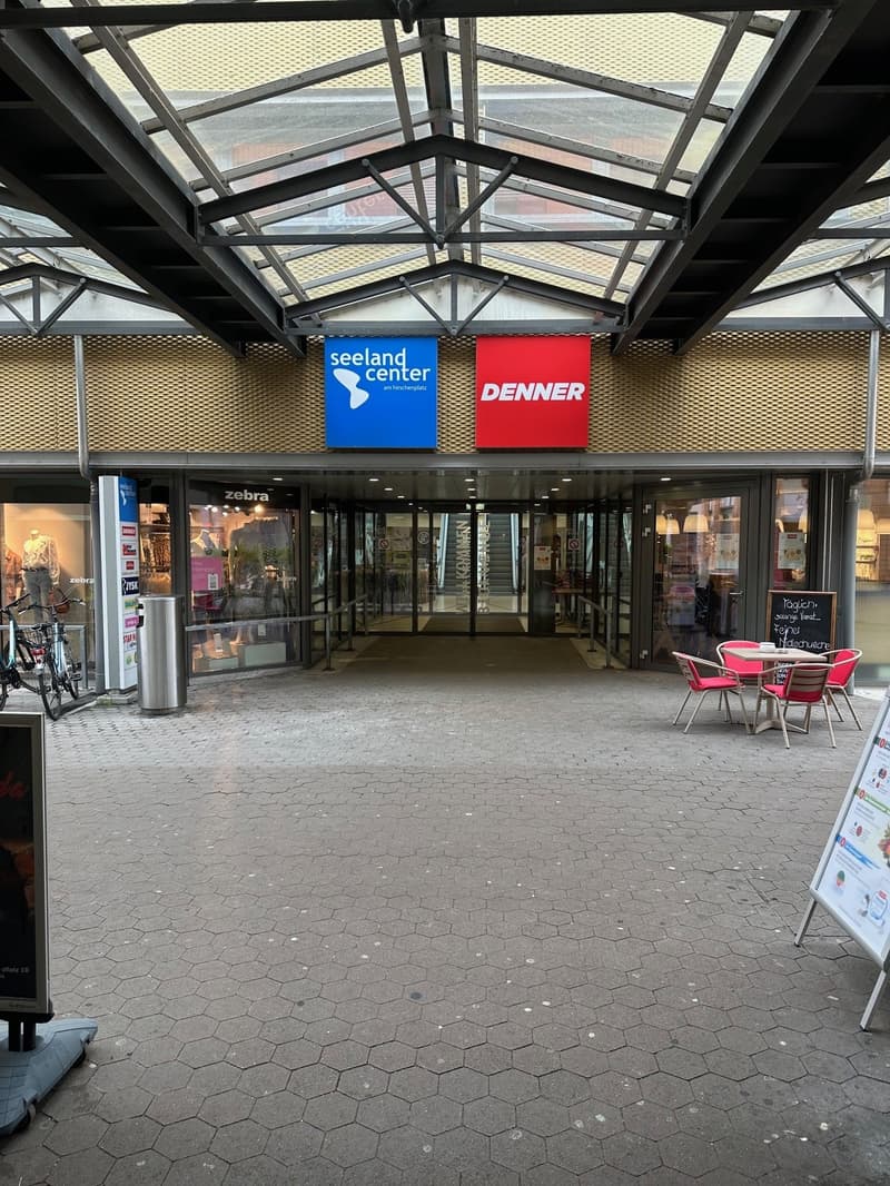 Entrance to a shopping center with glass doors, bicycles parked, a table and chairs outside, and a blackboard with writing.