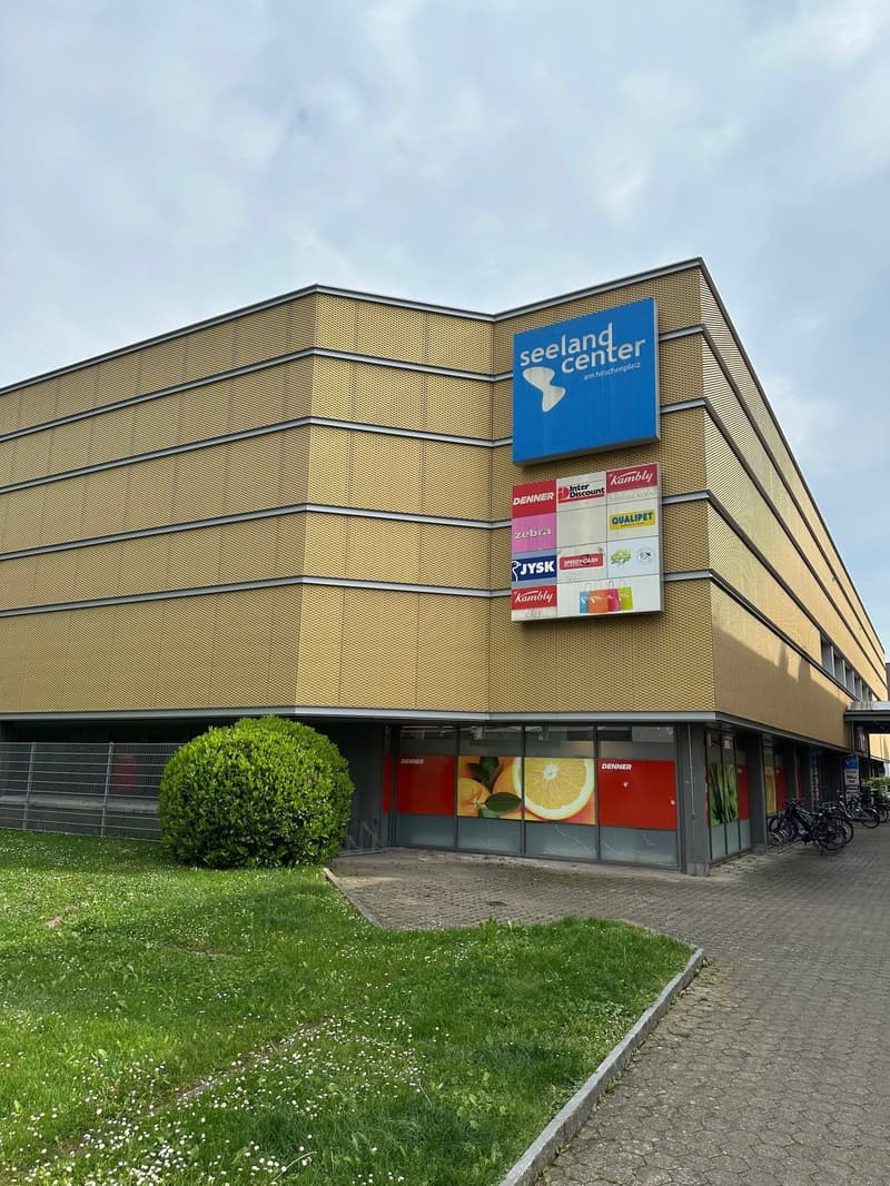A yellow building with a flat roof, glass windows, and a signboard with the text 'seeland center'. There are several advertisement boards attached to the wall. A few bicycles are parked in front of the building. A green bush is in front of the building, a