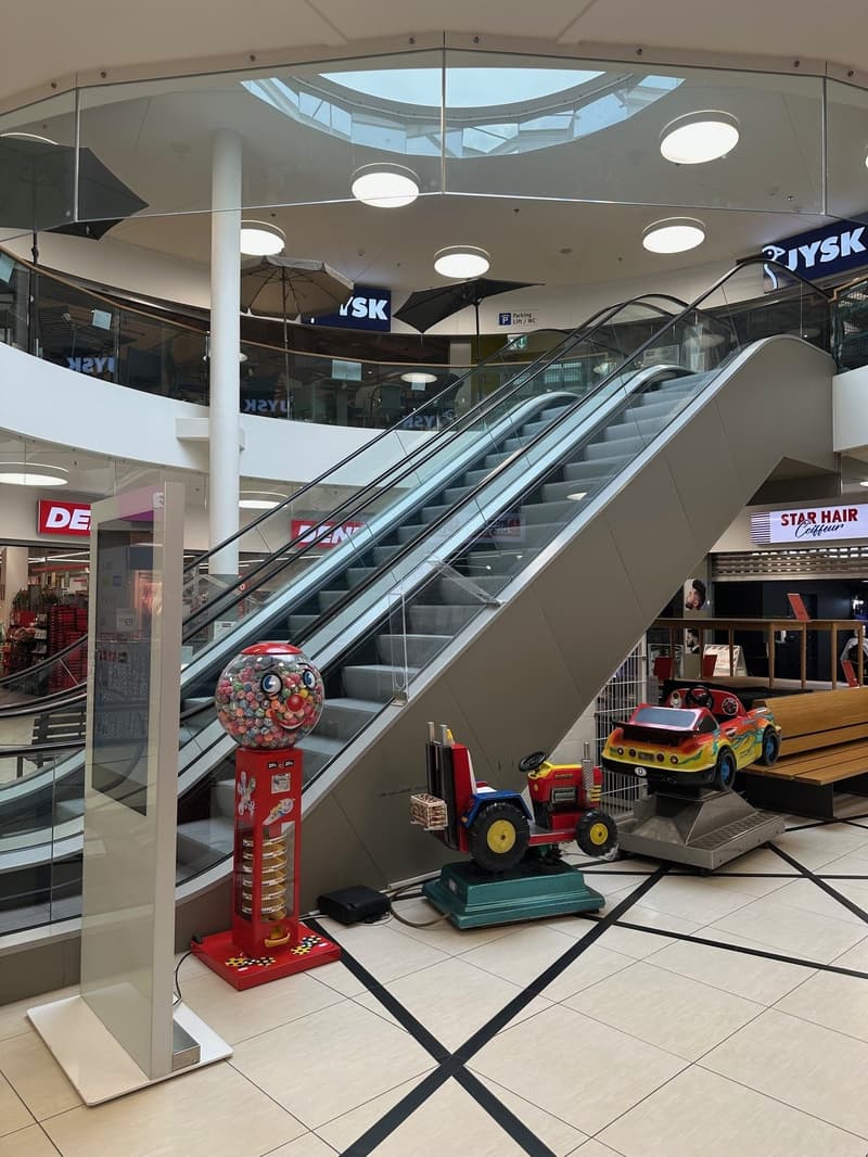 Mall interior with escalator, gumball machine, toy cars, and tiled floor