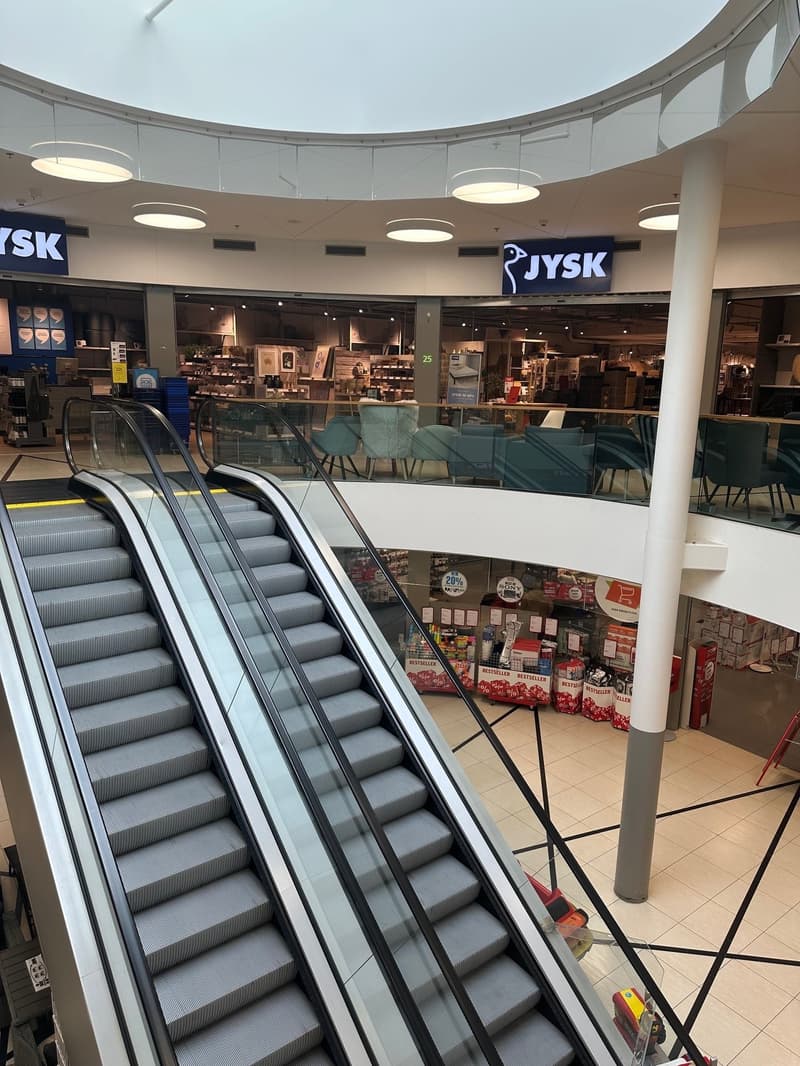 Mall interior with escalators, glass railings, and stores on the upper level