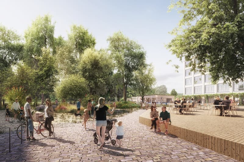 A paved walkway with people walking and cycling, tables and chairs for sitting, trees and plants, a building in the background, a playground with a water feature