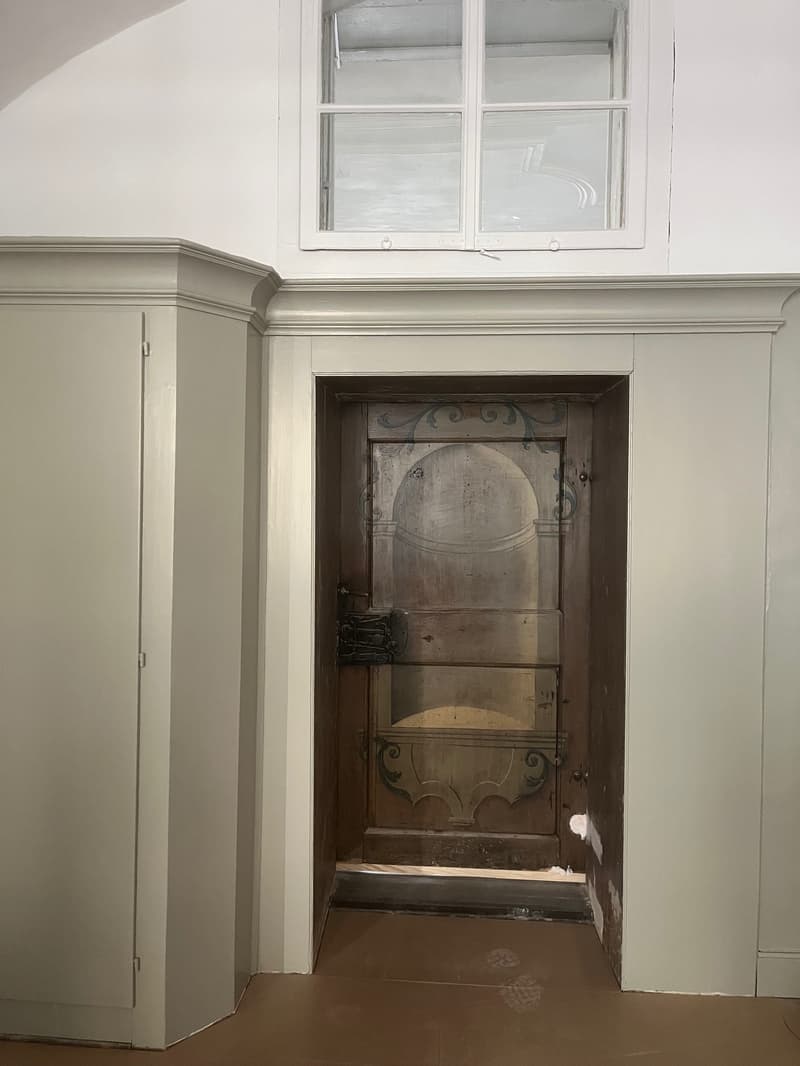 white walls, wooden door, small window, wooden floor